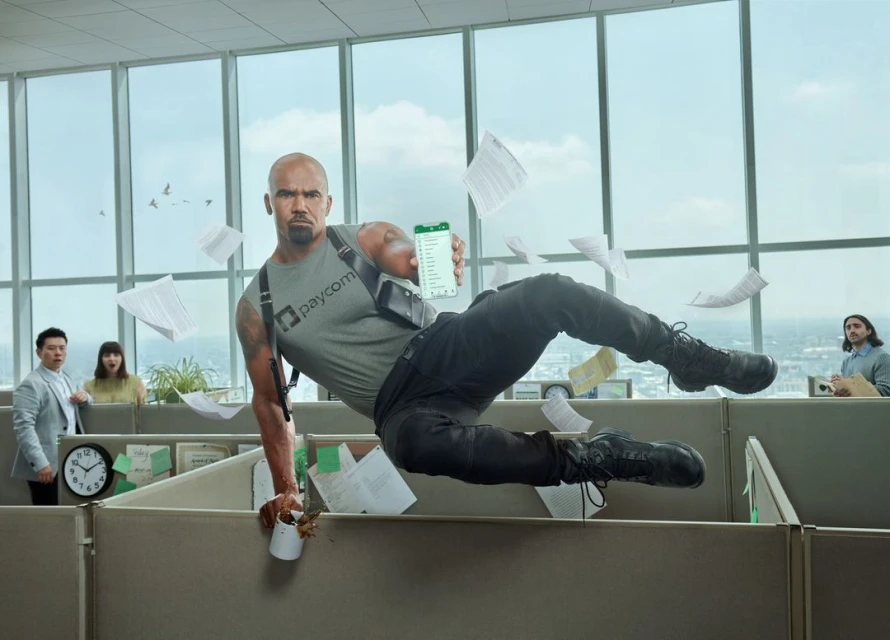 An adult male leaping over a cubicle in a modern office, holding a smartphone and wearing tactical gear, with surprised coworkers in background.